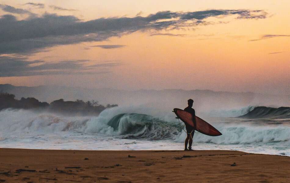 a man watching the big waves on a beach with his surfing board in a beautiful sunset time